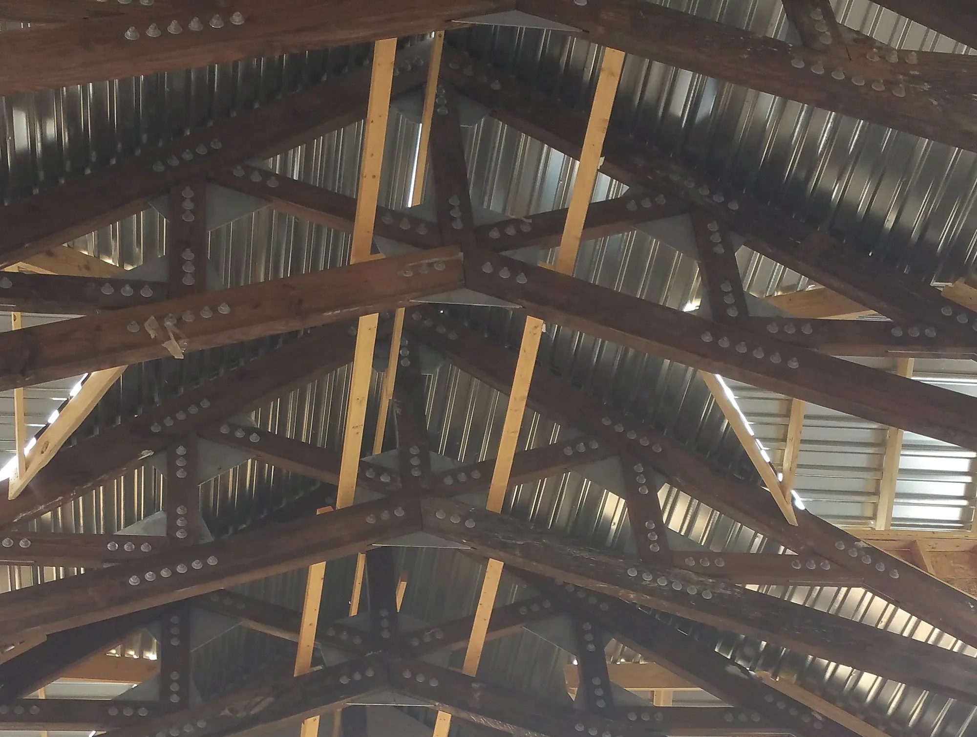 Looking up at dark stained timber trusses crossing in a repeating pattern, rows of bolts at every joint and steel gusset plates, with light wood blocking against a corrugated metal roof deck.