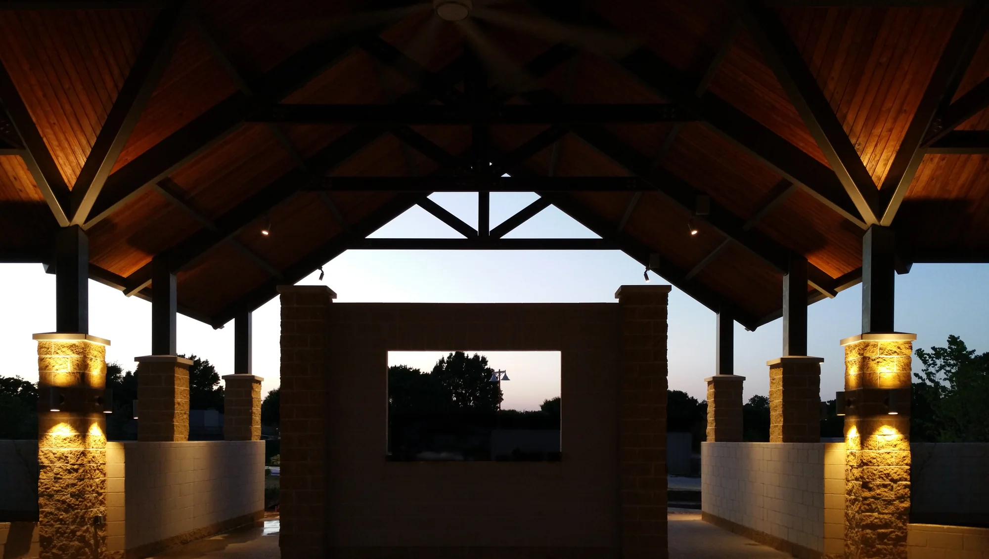 Symmetric view through an open pavilion at dusk, warm lights washing the rough-faced block masonry columns and wood ceiling, a central block wall with a window opening framing trees against a fading sky.