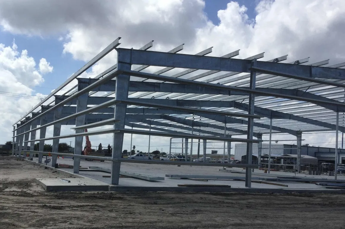 Galvanized steel columns, rafters and purlins of a pre-engineered metal building rise from a fresh concrete slab under a blue sky with white clouds.