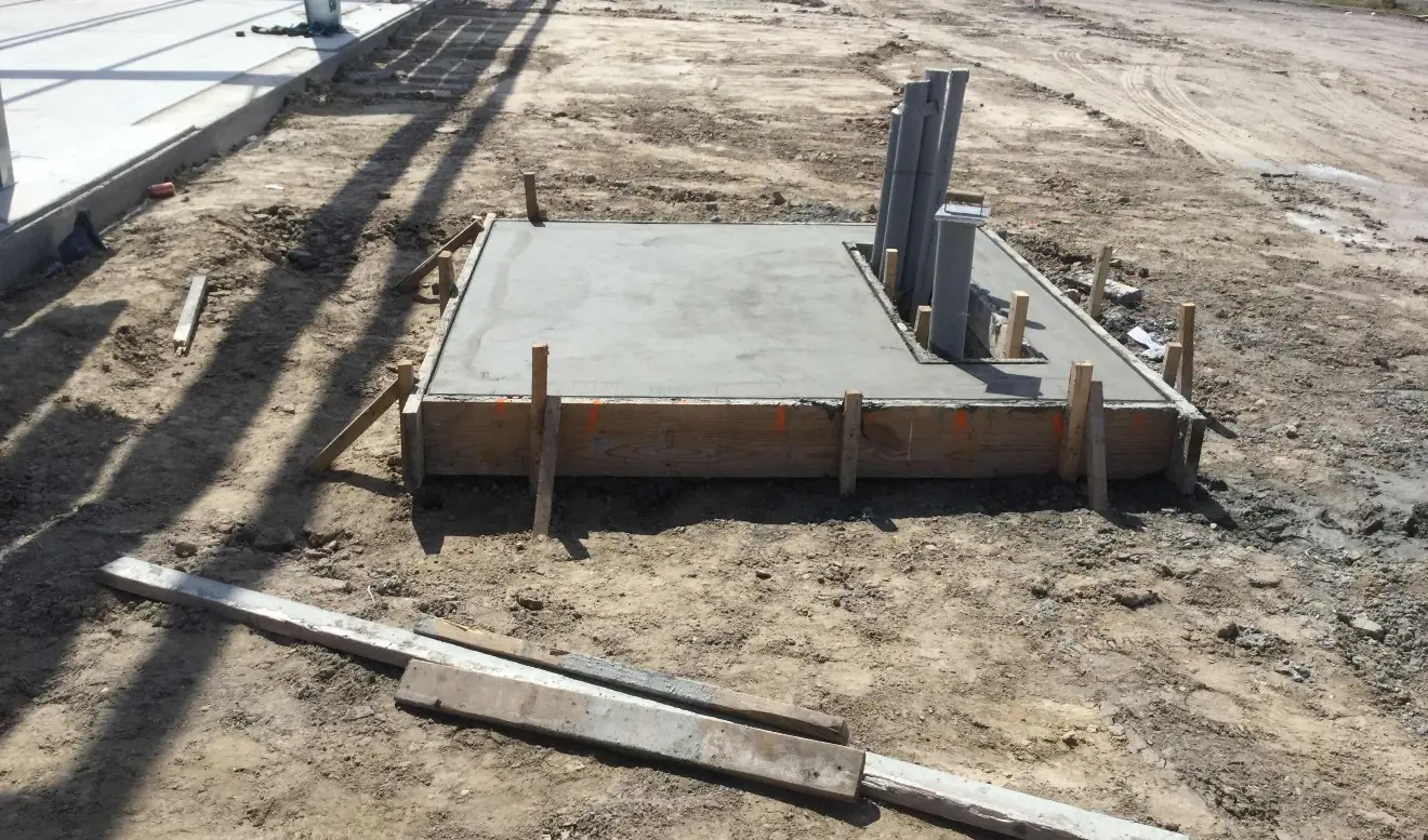 A small formed concrete pad sits on bare dirt with a cluster of grey conduits rising through a rectangular opening at its back corner, loose form boards in the foreground and long shadows of the steel frame across the ground.