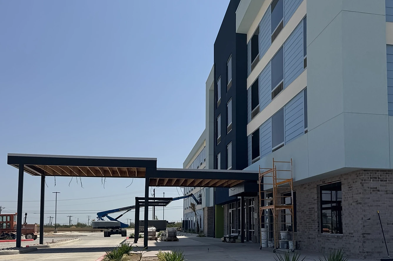 A four-story hotel with light blue lap siding, pale stucco and a plain navy stair tower running away from the camera, a steel entry canopy with open wood framing and loose light wires, a blue boom lift and pallets in the drive, new plantings behind a red curb, and a street sweeper at the far left under a clear sky.