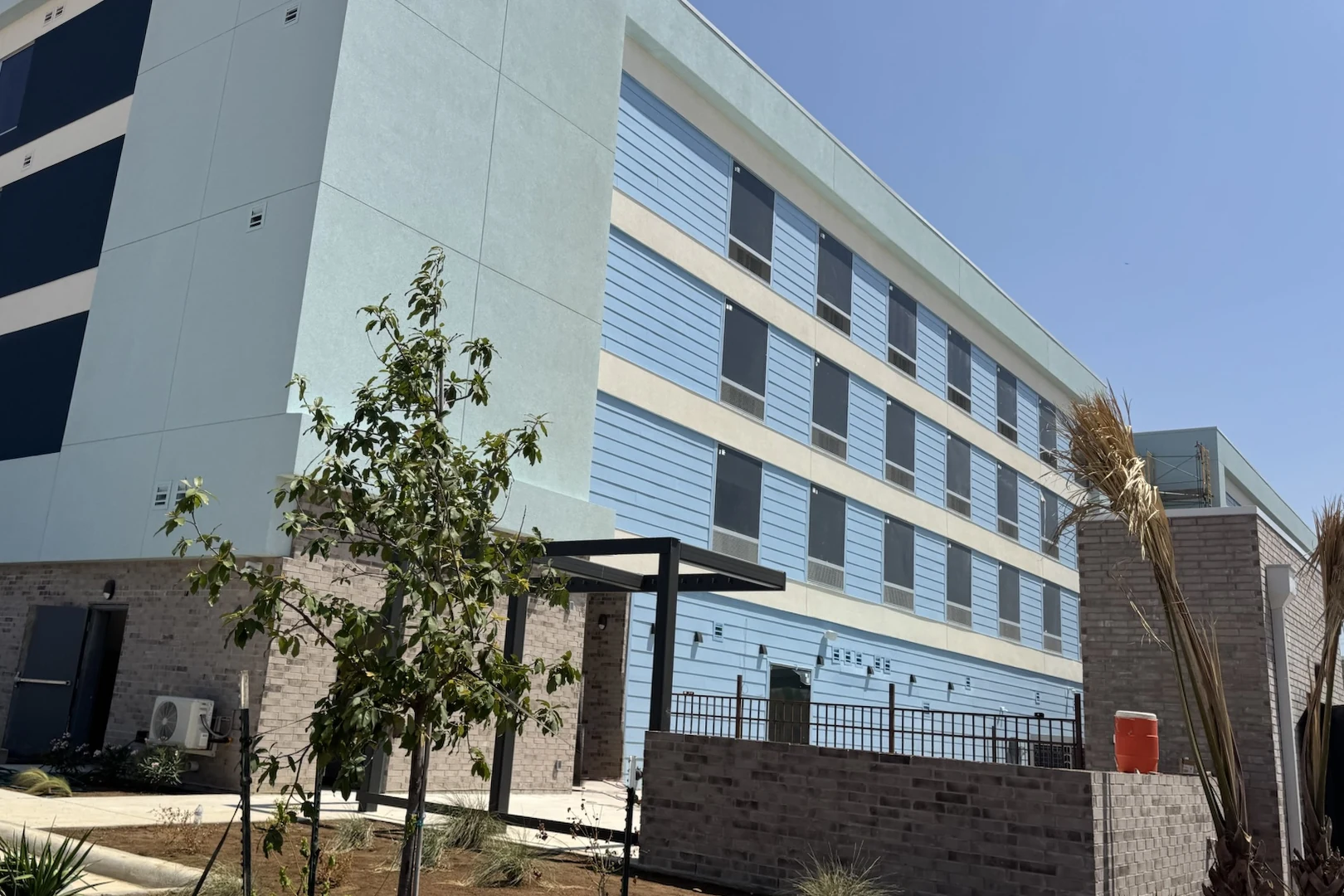 A four-story hotel seen from a low corner angle under a clear blue sky: a pale mint stucco tower with navy window panels at left, a long light blue lap-sided wing with four rows of windows running off to the right, a gray brick base with a service door and a small wall-mounted AC unit, a black steel shade frame at a side entry, a low brick wall topped by a metal fence, a young tree and fresh planting beds in front, and an orange water jug on the wall at right.