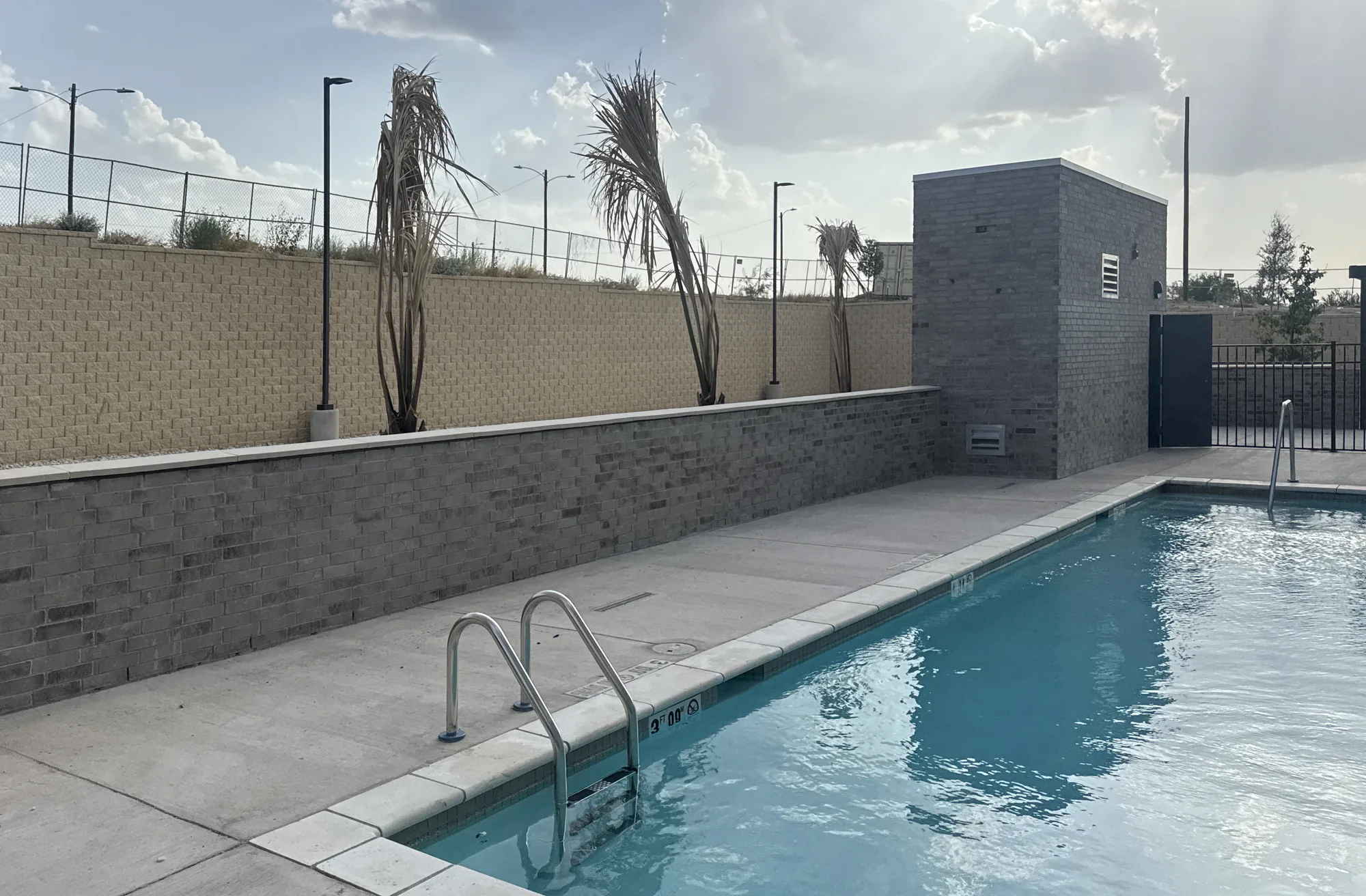 A filled outdoor pool with a stainless ladder and depth marker, a concrete deck, a gray brick planter wall and a tan retaining wall behind, and a small brick equipment building, under a partly cloudy sky.
