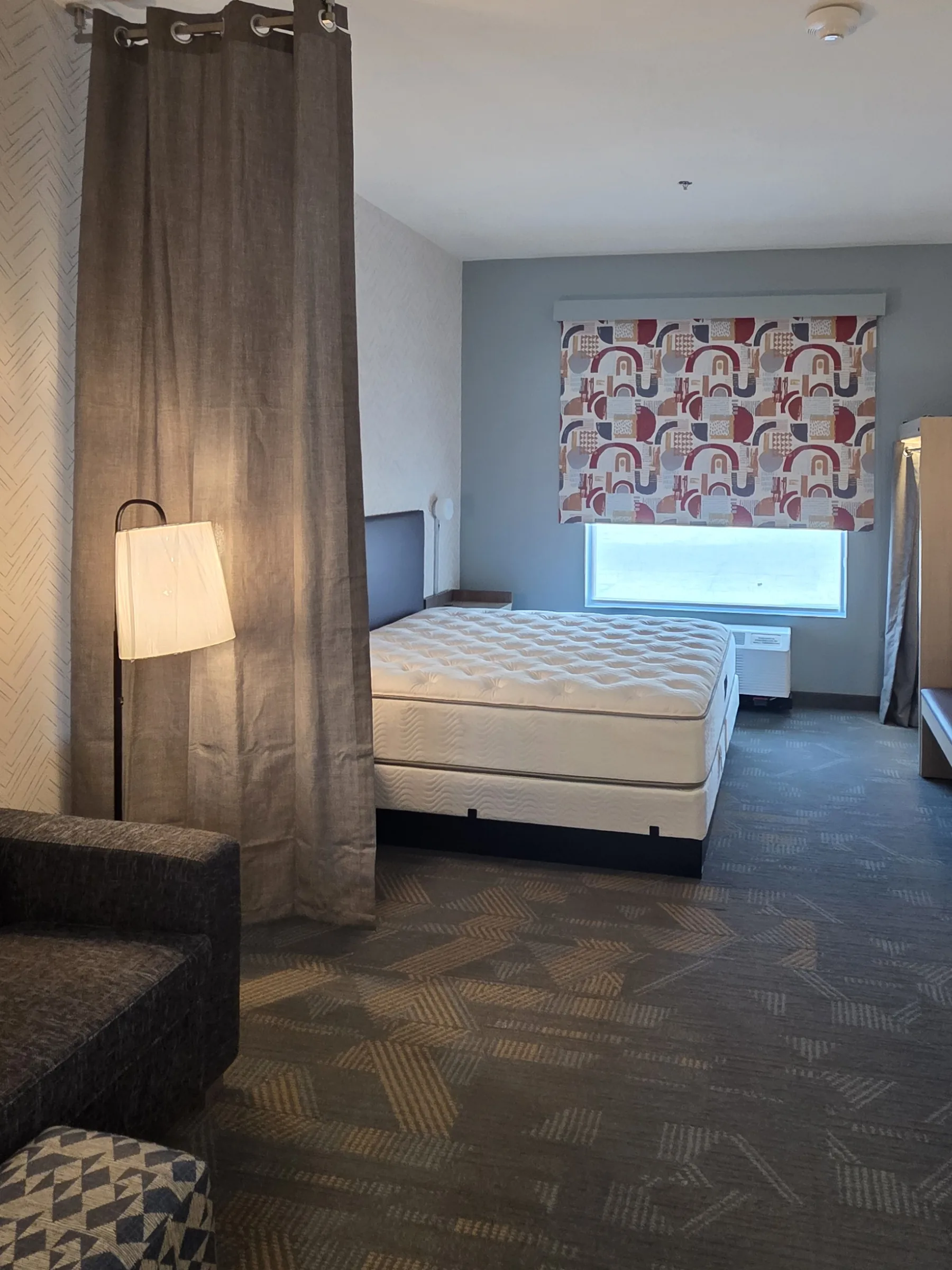 A furnished hotel guest room: a gray curtain divider and floor lamp at left, a bare king mattress, a window with a patterned shade on a blue-gray wall, an open closet at right, and a gray sofa and patterned ottoman in the foreground.