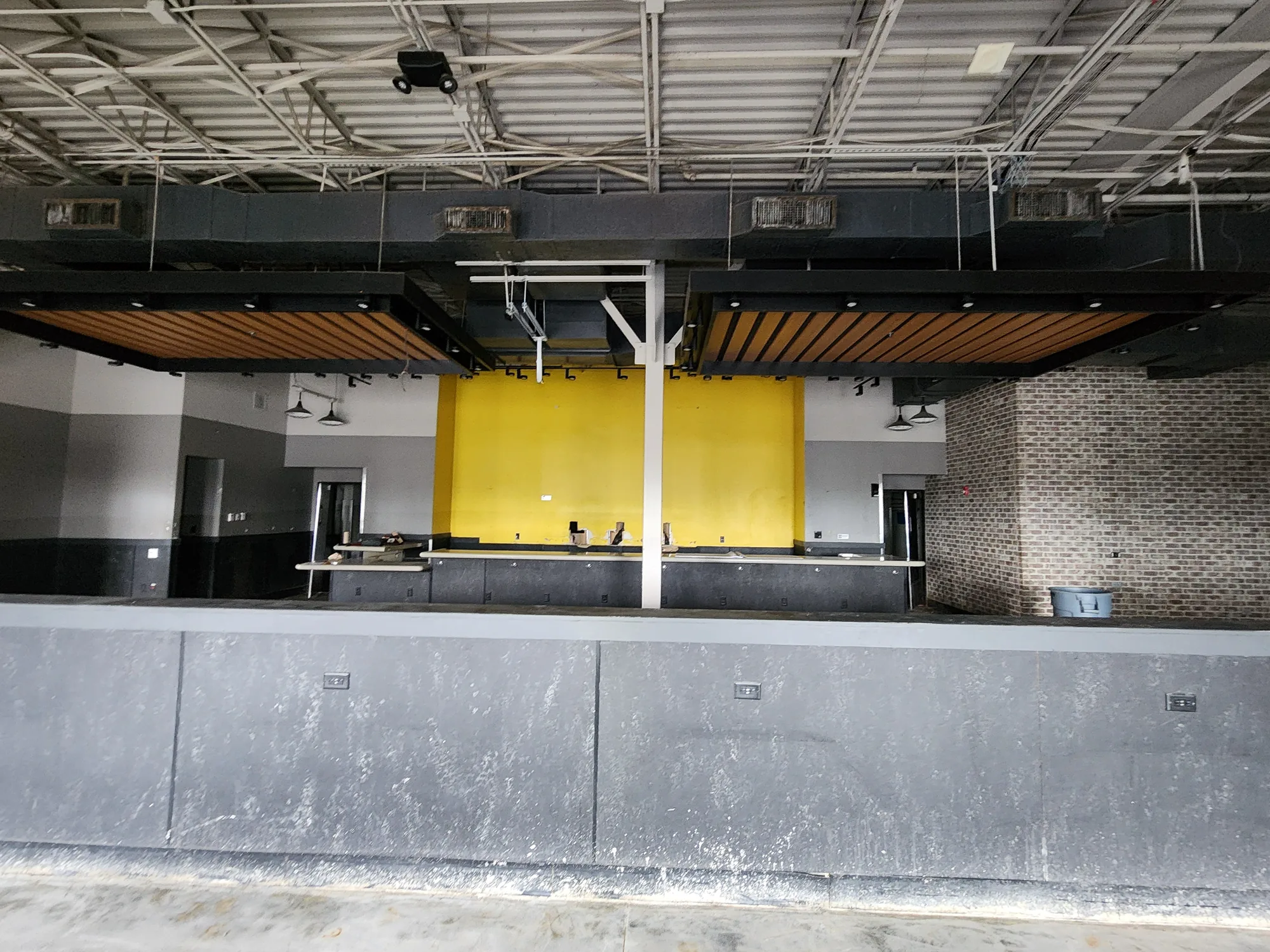 Before: Long gray concrete bar counter in front of a yellow accent wall, a brick wall, and wood slat soffits under an open ceiling with exposed ductwork.