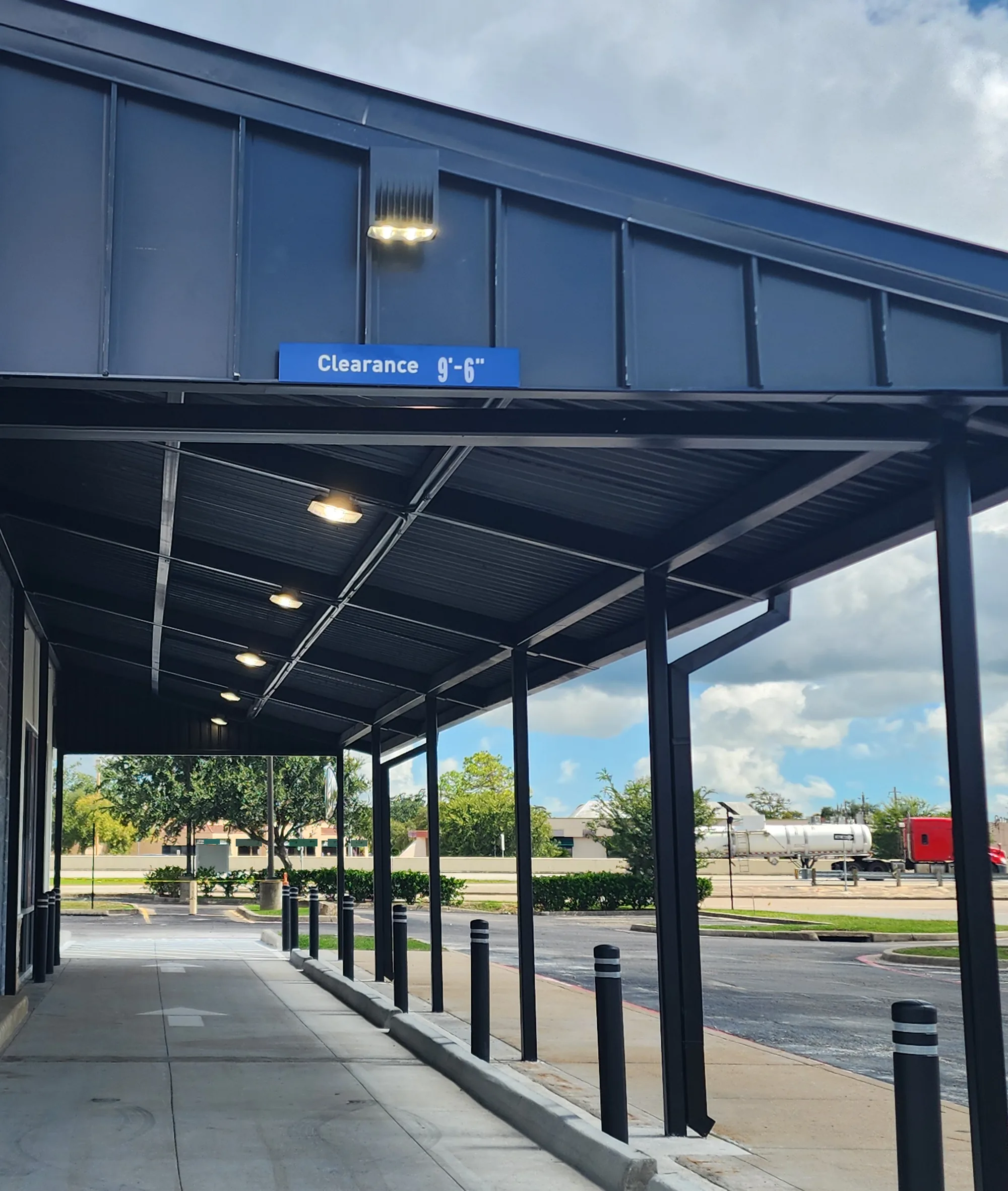 View along a covered drive-through lane with a dark metal canopy, round ceiling lights, black bollards, and trees, buildings and a passing tanker truck beyond.