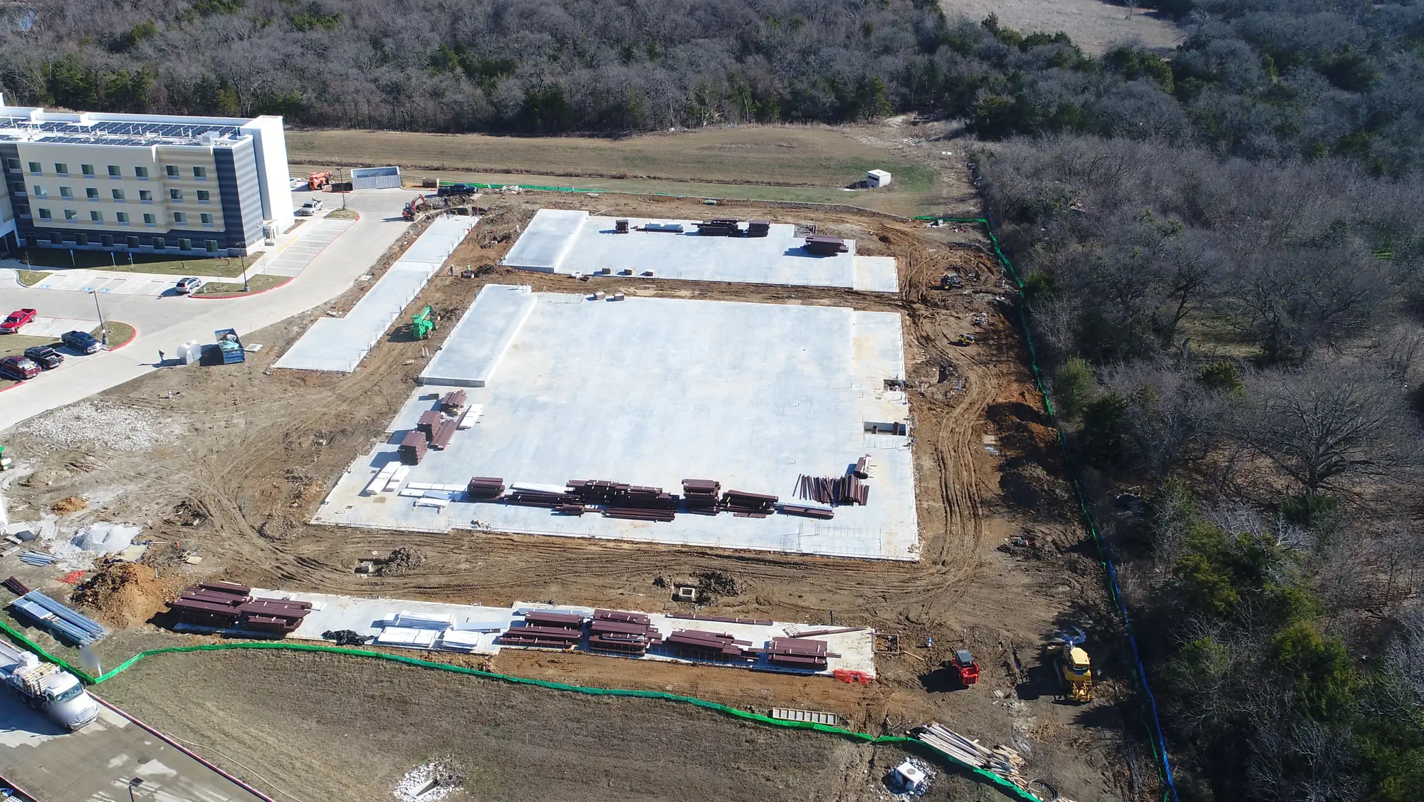 Aerial view of three poured concrete slabs on a cleared site edged by green silt fence, stacks of red-primed steel beams laid out on and beside them, a flatbed truck at lower left, part of a hotel and its parking lot at upper left and bare winter woods at right.
