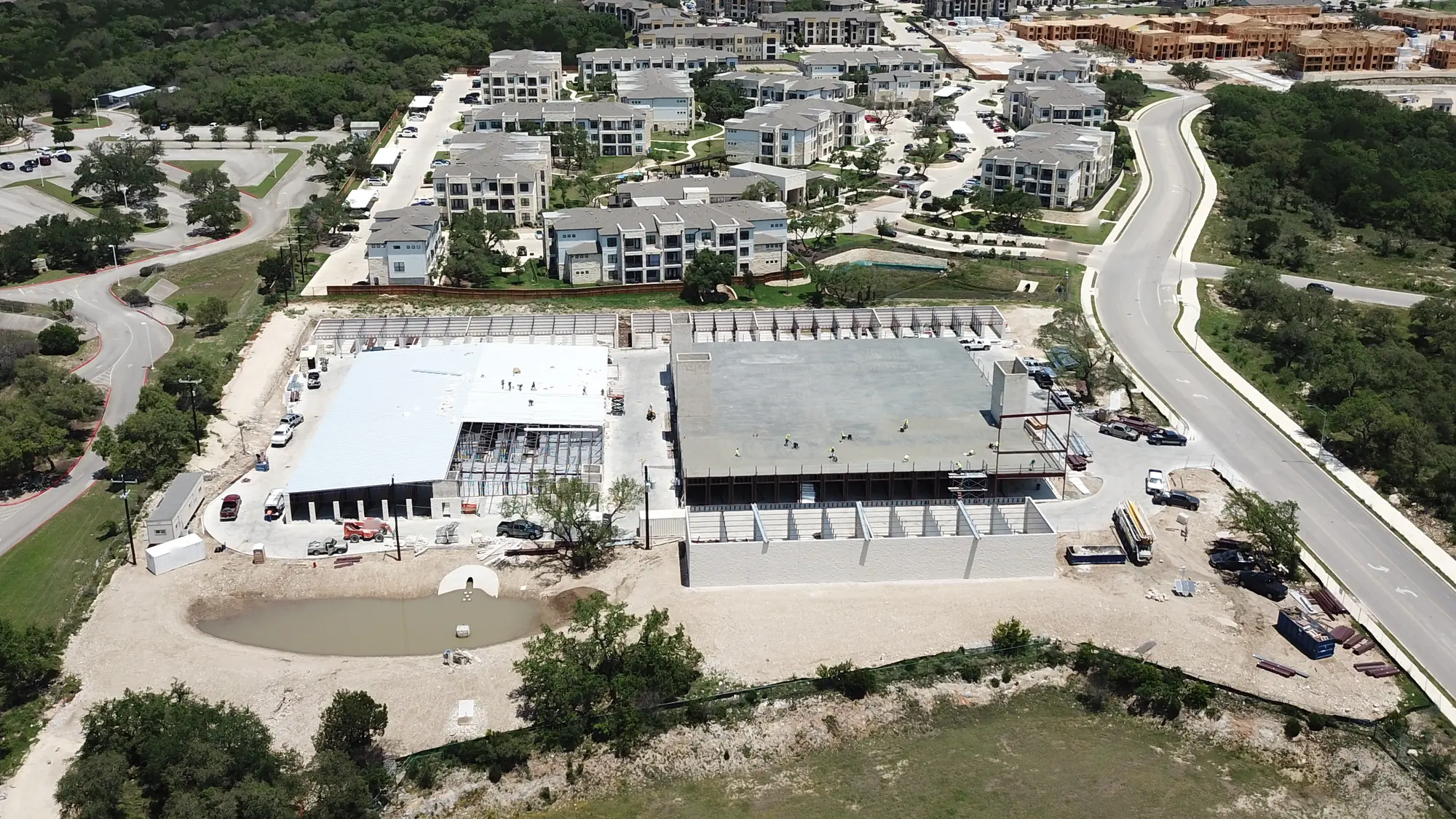 Aerial view of the same site eight months later: crews on a gray concrete deck set on one story of steel, with stair and elevator shafts rising above it, a white-roofed building at left with open framing at one end, rows of drive-up framing, trucks, lifts and a detention pond in front, and the apartments beyond.
