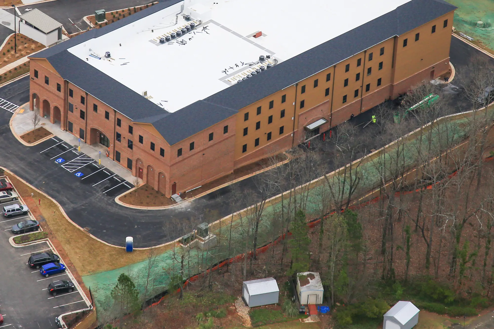 Angled aerial of a three-story brick and tan storage building with a white roof and dark shingled mansard edge, arched ground-floor openings at the front, fresh paving, green hydroseed on the slopes and bare winter trees around it.