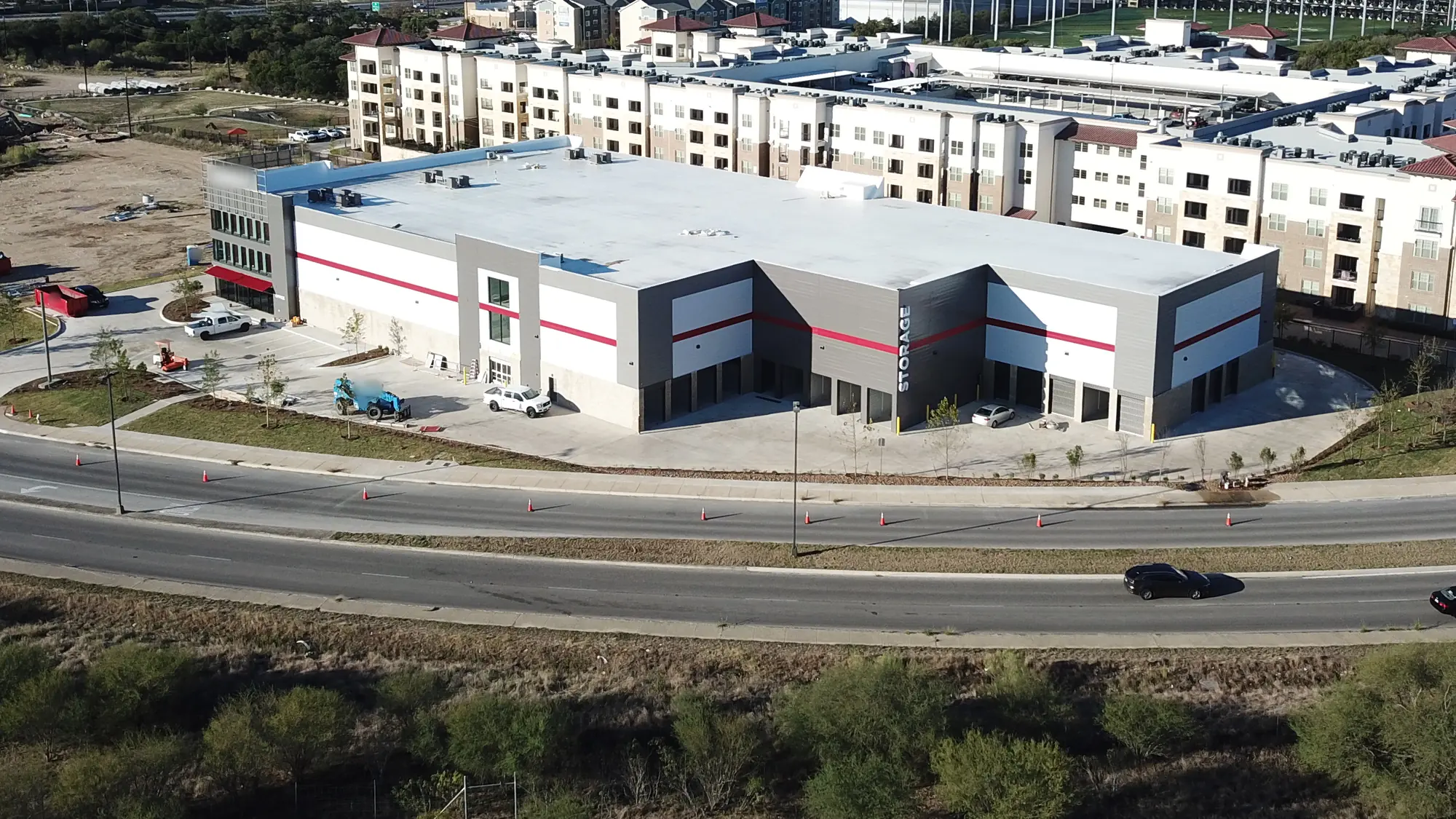 Angled aerial of a white and gray storage building with a red accent stripe, a vertical STORAGE sign and a glass corner tower with a red awning, a lift and pickups in the front lot, a curving four-lane road with orange cones in front, scrub brush in the foreground and large apartment buildings behind.