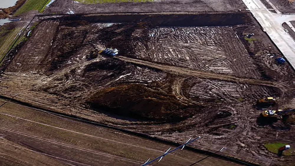Aerial view of a muddy cleared site with tire-tracked dirt and a dug-out area in the middle, two yellow excavators at right and a dozer at top center, the new concrete street at upper right, the pond edge at upper left and a power line across the front.