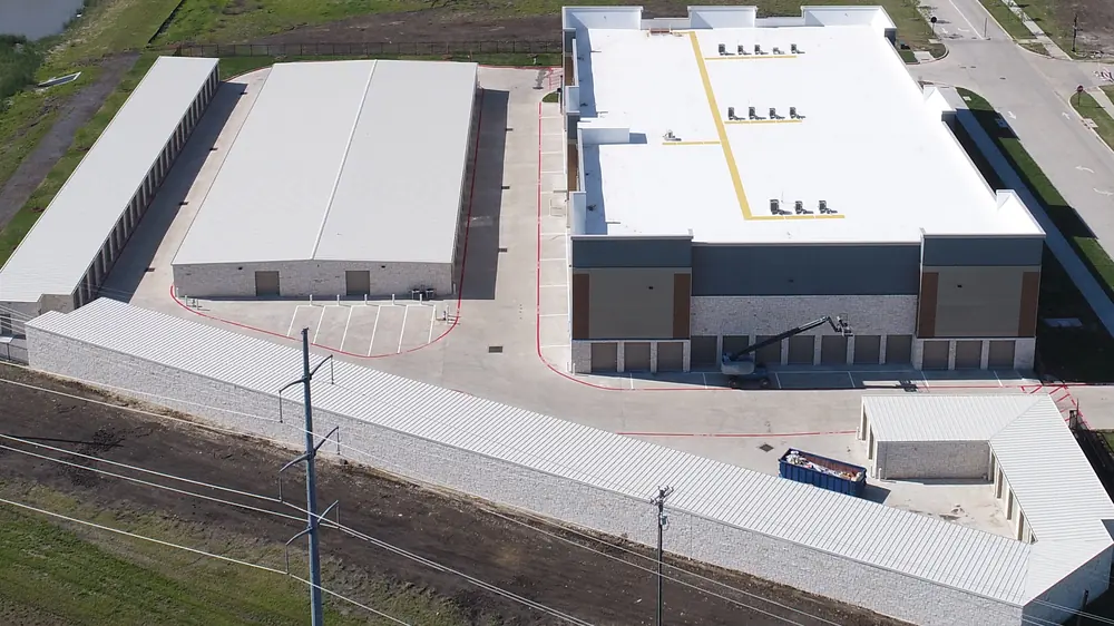 Aerial view of the facility near completion: a large white-roofed main building with brown and gray panels and a row of roll-up doors, long single-story drive-up buildings with white metal roofs, new paving, a boom lift at the main building and a loaded dumpster in the drive, the pond at upper left, the new street at upper right and the power line across the front.